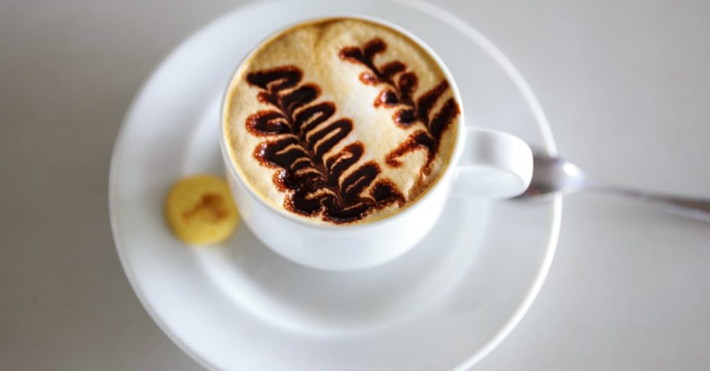 Close-up of a cappuccino with intricate latte art, served on a white saucer with a small biscuit.